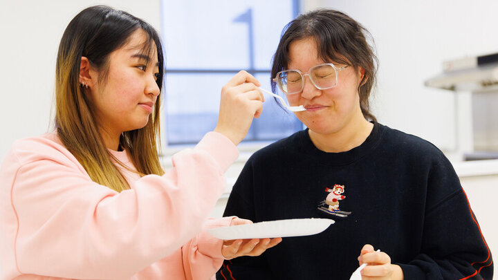 Two young adults, one is feeding the other off of a paper plate. 
